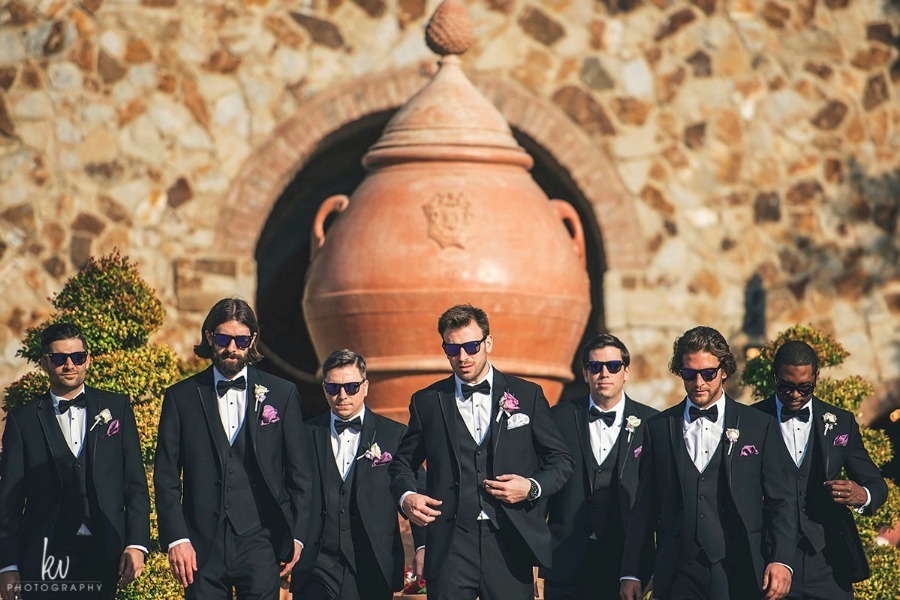 Groom and groomsmen in black tuxedos outside the stone Clubhouse at Bella Collina