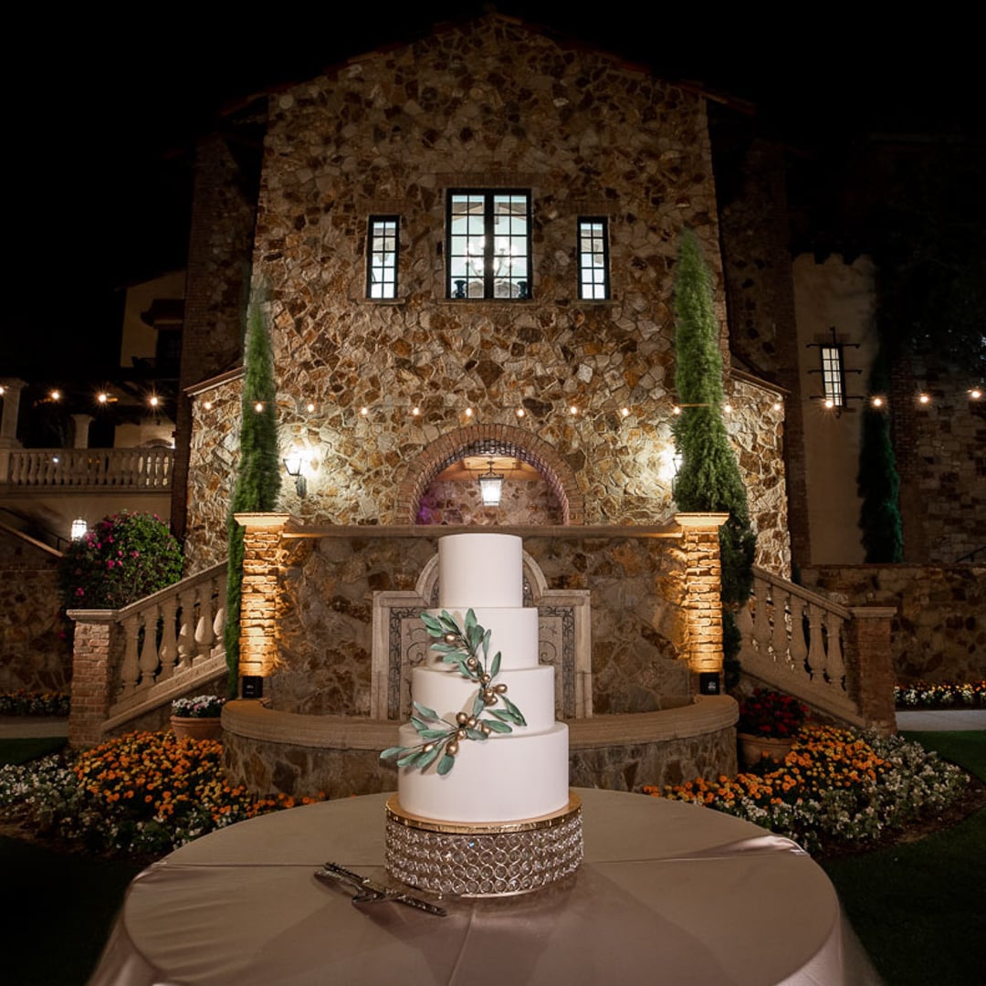 White wedding cake with greenery displayed in front of the illuminated Bella Collina Clubhouse