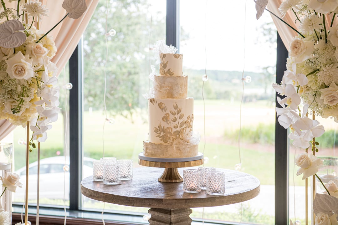 Elegant three-tier wedding cake with gold floral details framed by white flowers at Bella Collina