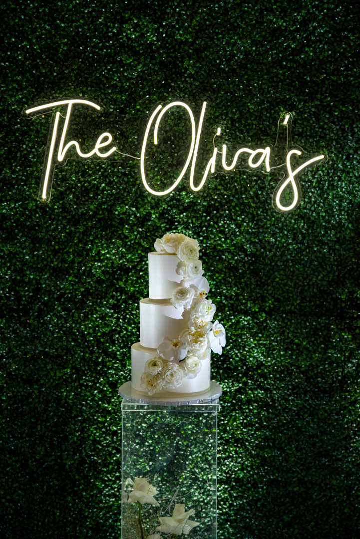 White and gold wedding cake displayed against a greenery wall with a neon sign at Bella Collina