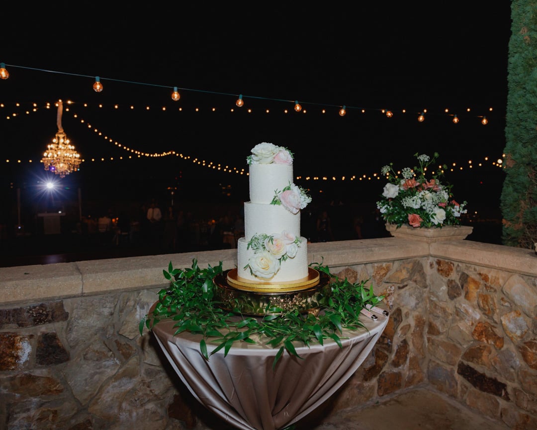 Three-tier white wedding cake with greenery displayed under string lights at Bella Collina