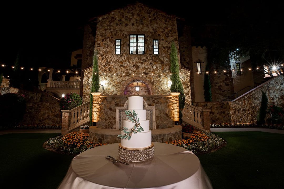 White wedding cake displayed in front of the illuminated stone Clubhouse at Bella Collina